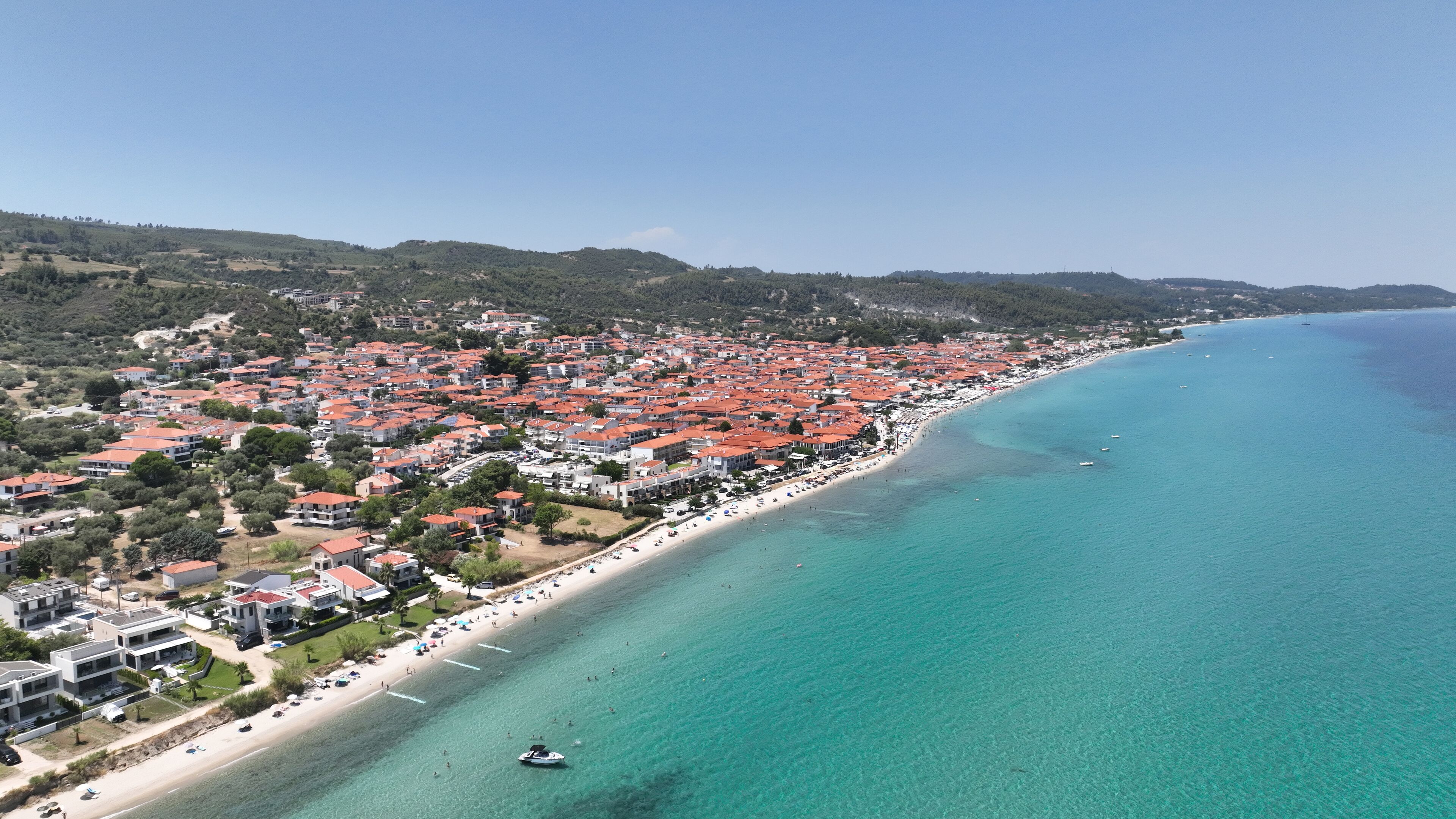 An aerial view of a coastal town with a long beach, red-roofed buildings, and clear blue waters.