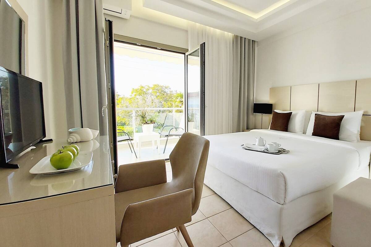 A hotel room with a bed, bedside tables, a TV, a desk with apples, a chair, and a balcony with a view.