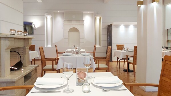 Comedor con mesas dispuestas para cuatro, cada una con mantel blanco, platos, cubiertos y copas. Hay una chimenea con repisa y elementos decorativos. La habitación tiene sillas de madera y un techo alto con iluminación empotrada.