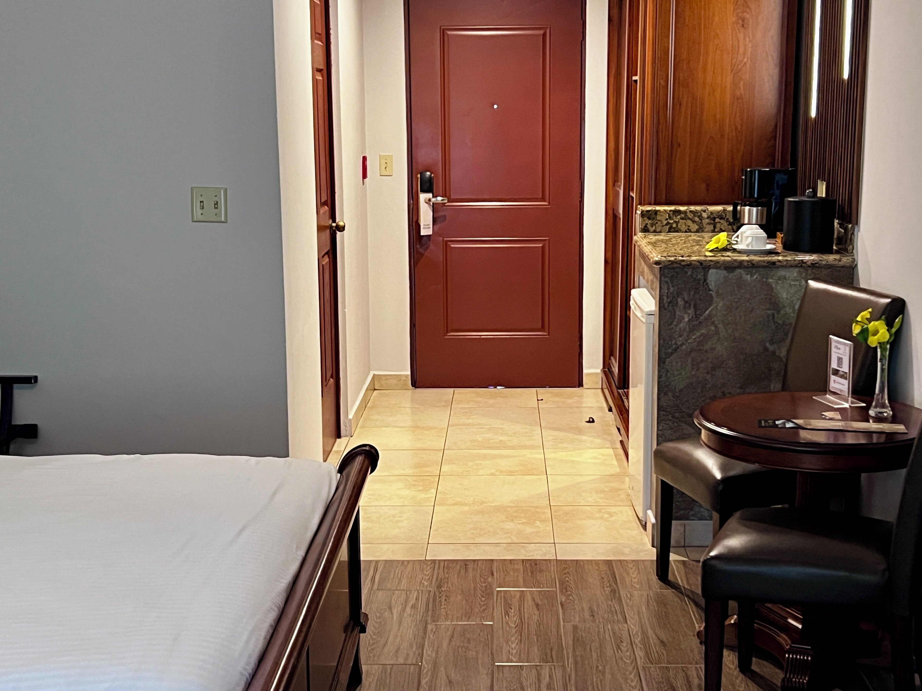 A hotel room with a bed, a wooden chair, a small table with a vase, and a kitchen area with a coffee machine.