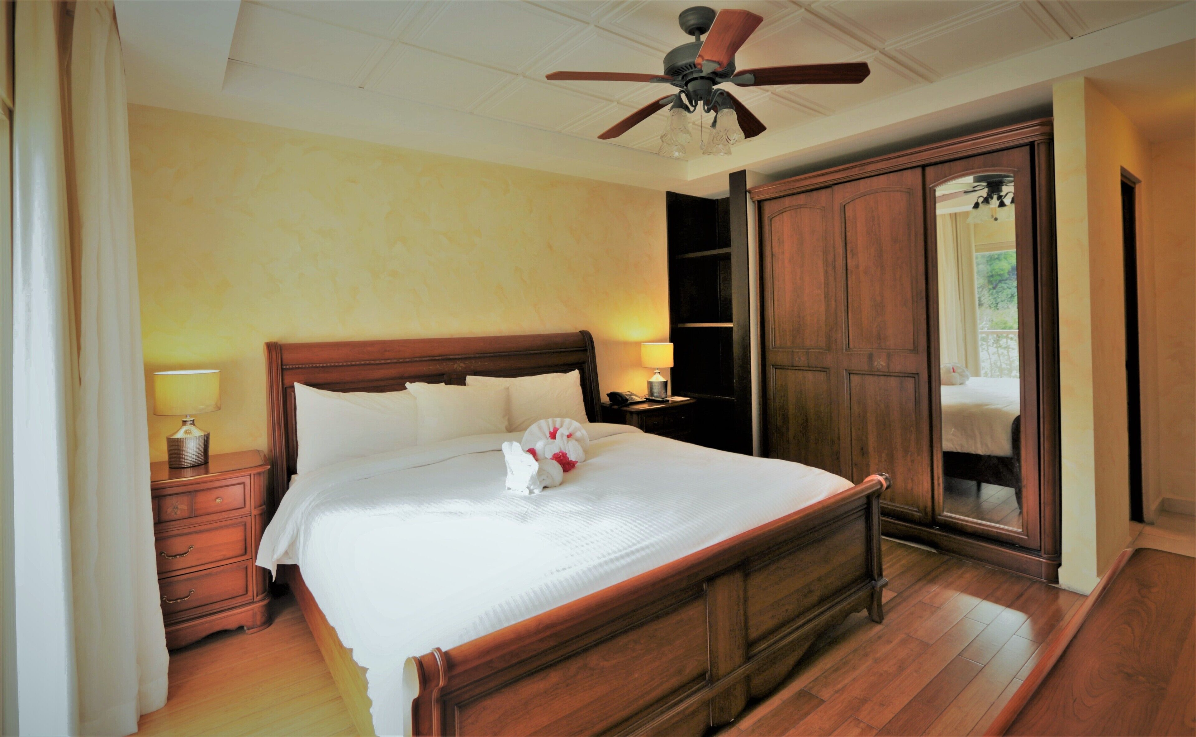 A neatly made bed with white linens, a wooden headboard, and a bedside lamp. There is a wooden wardrobe with a mirror and a ceiling fan above the bed.