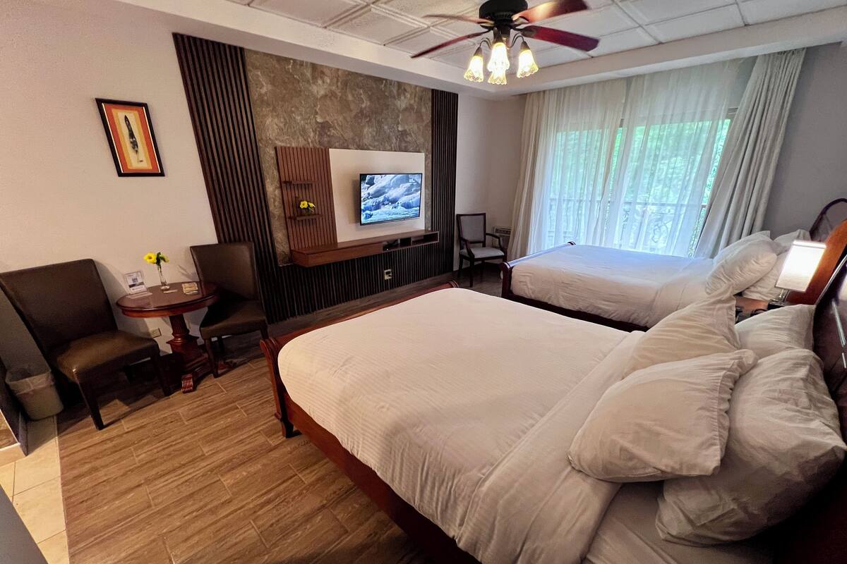 A hotel room with a large bed, a smaller bed, a desk with a chair, a TV, and a ceiling fan.