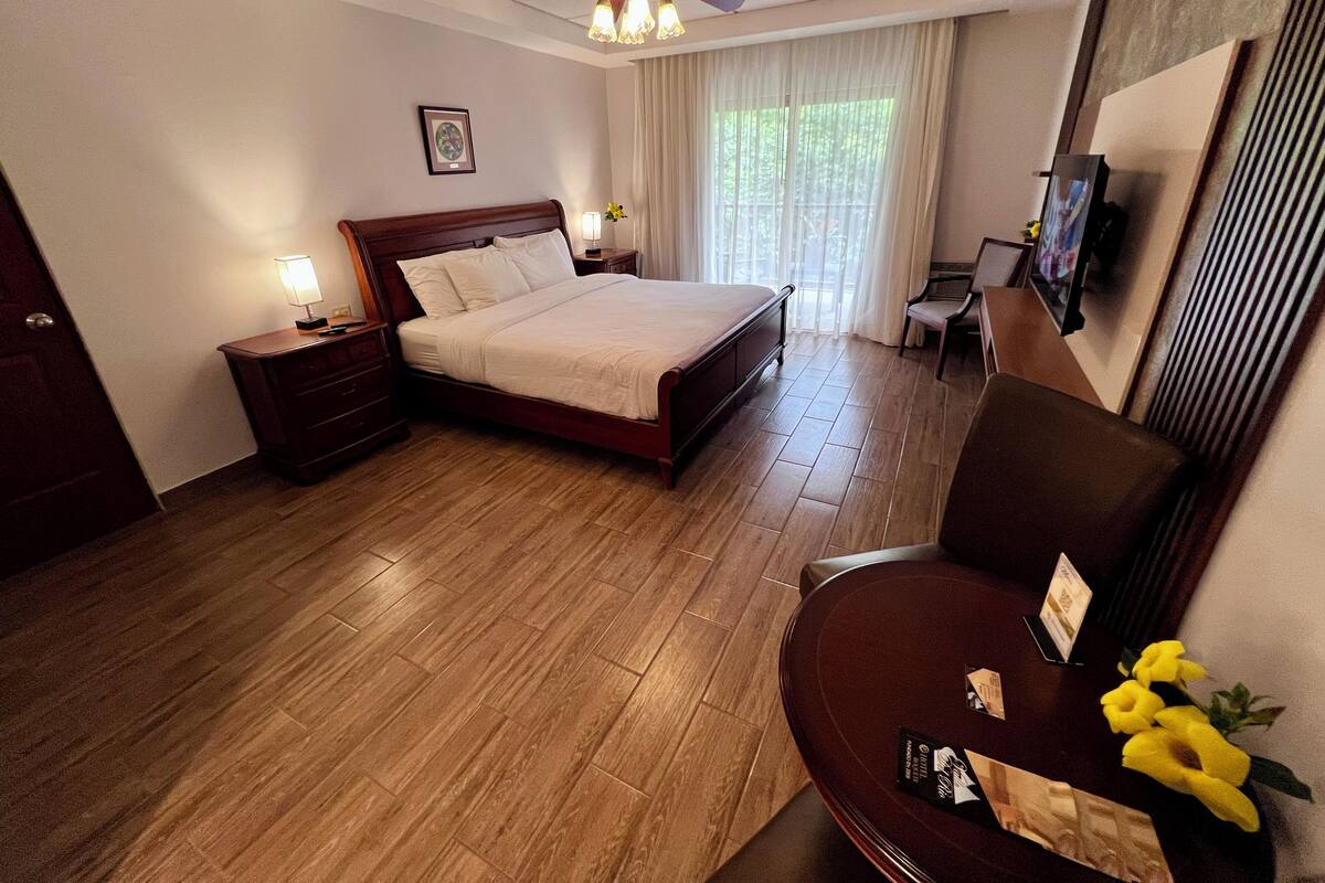 A hotel bedroom with a wooden floor, a bed with white linens, a nightstand, a desk with a chair, a ceiling fan, and a window with curtains.