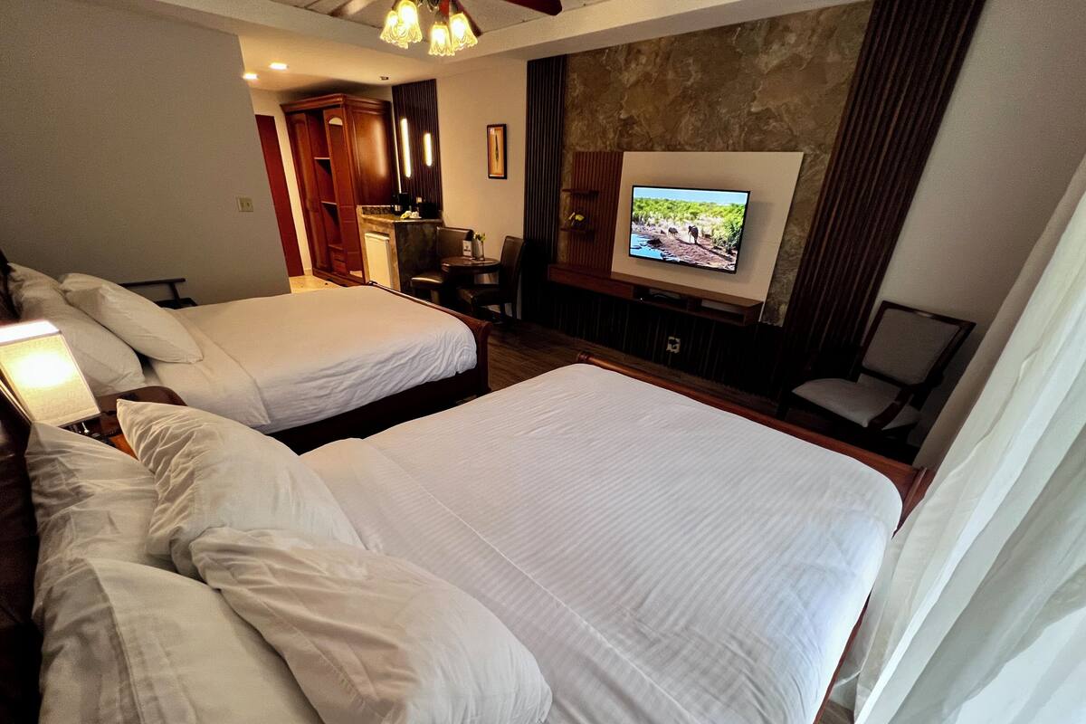 A hotel room with two large beds, a television mounted on the wall, a desk with a chair, and a small seating area with a chair.
