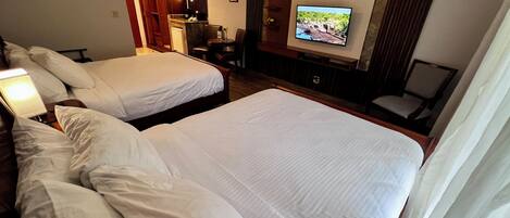A hotel room with two large beds, a television mounted on the wall, a desk with a chair, and a small seating area with a chair.