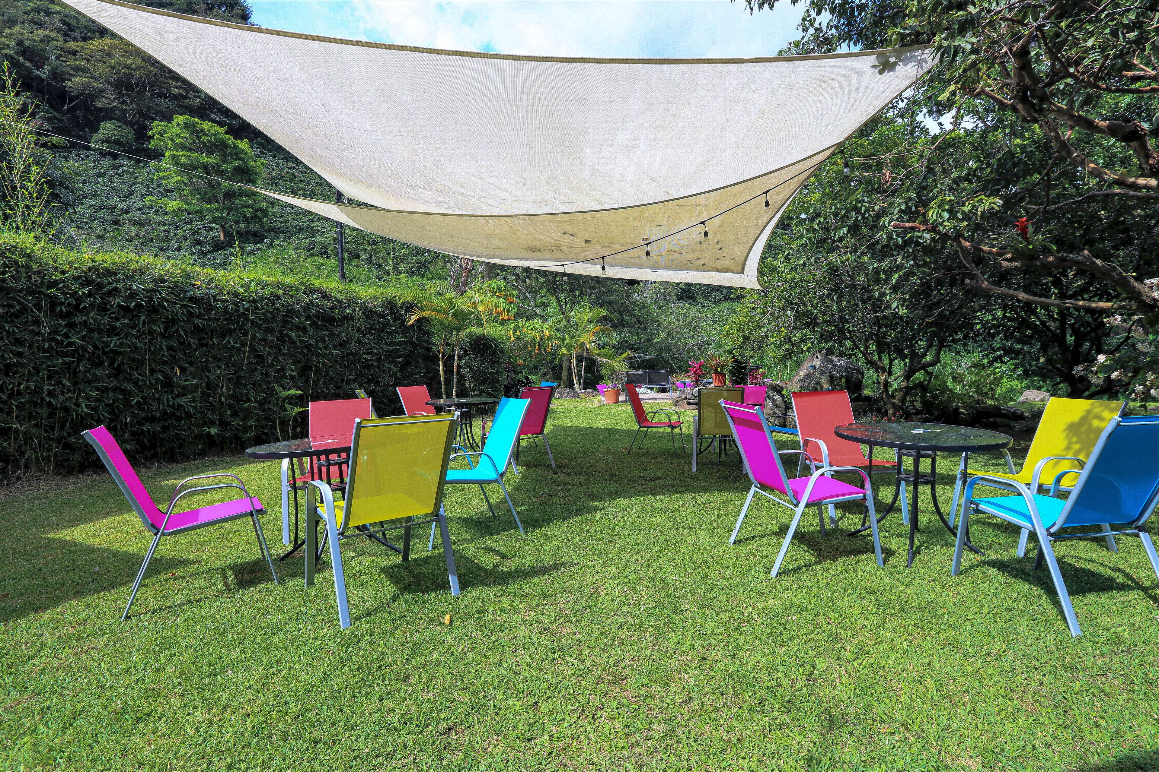 Un área de estar al aire libre con sillas y mesas de colores bajo una vela de sombra.