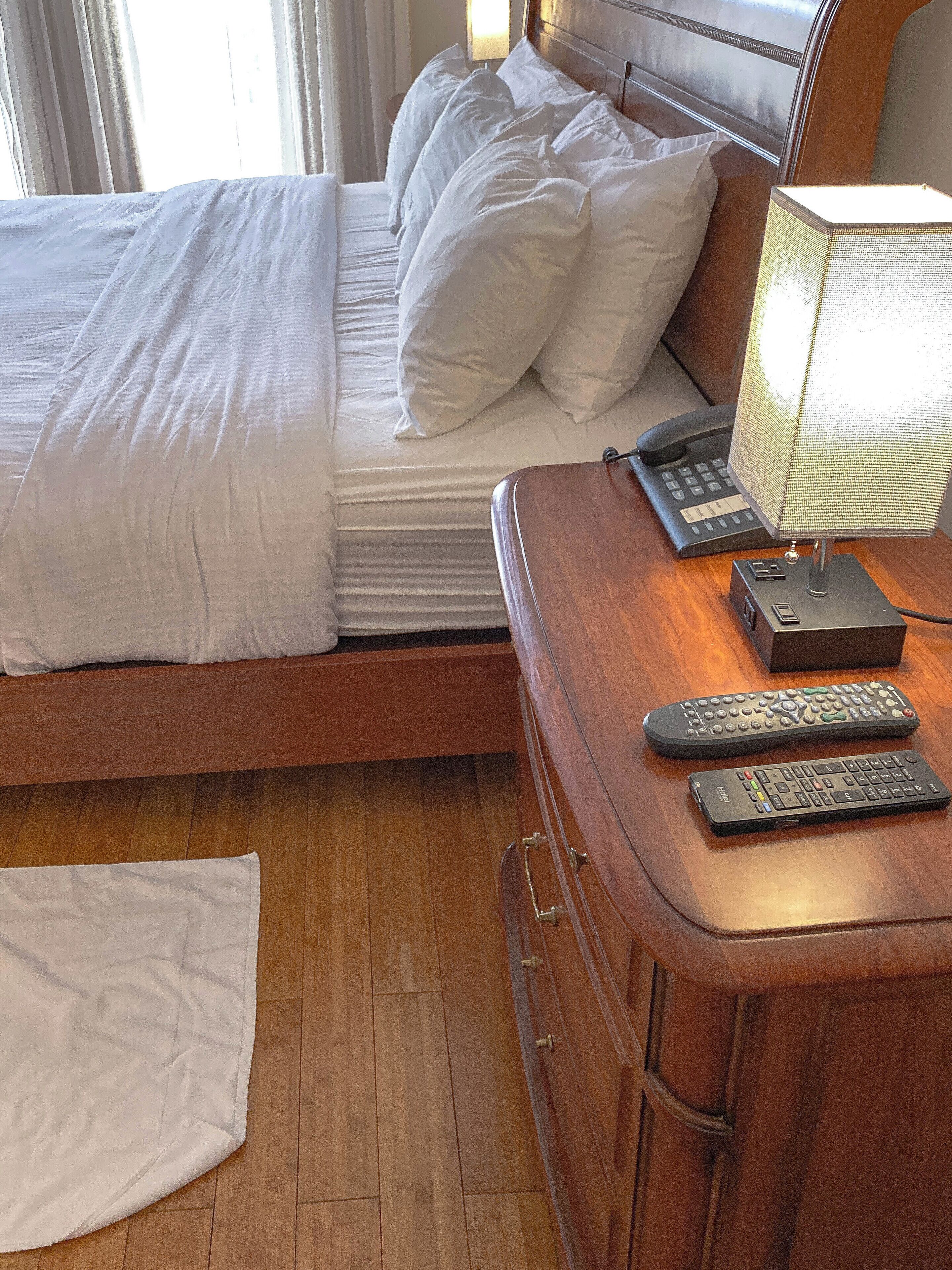 A hotel room with a bed, a desk with a lamp and a telephone, and two remote controls on the desk.