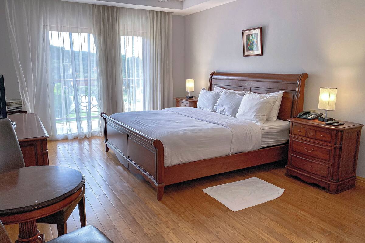 A hotel room with a wooden bed, a desk, a chair, and a view of the outdoors through large windows.