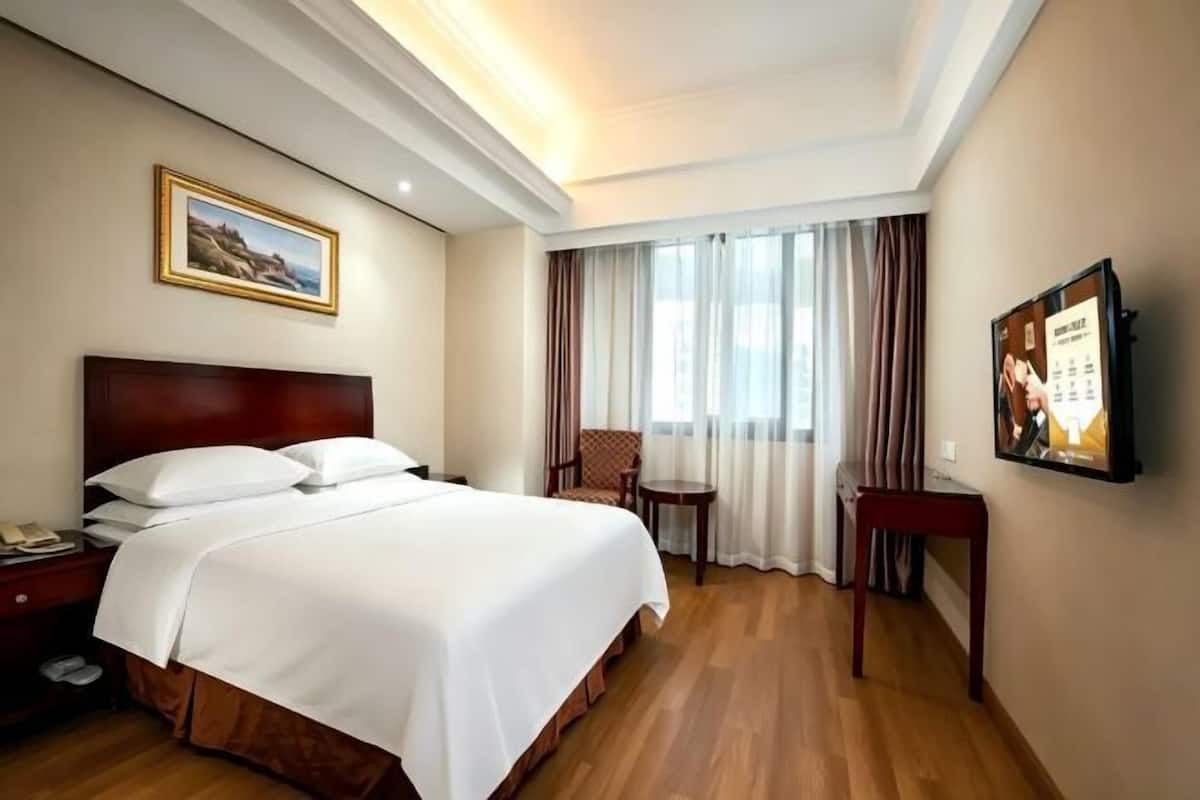 A hotel room with a large bed, a desk, a chair, a small table, a framed picture on the wall, and a window with curtains.