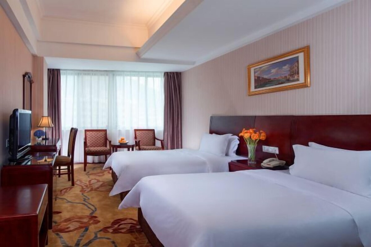 A hotel room with two beds, a desk with a lamp, a chair, a TV, and a painting on the wall.