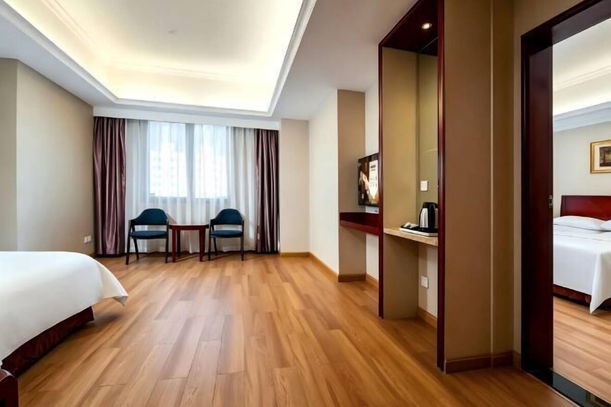 A hotel room with a bed, two chairs, a small table, a desk, a mirror, and a view of another room.