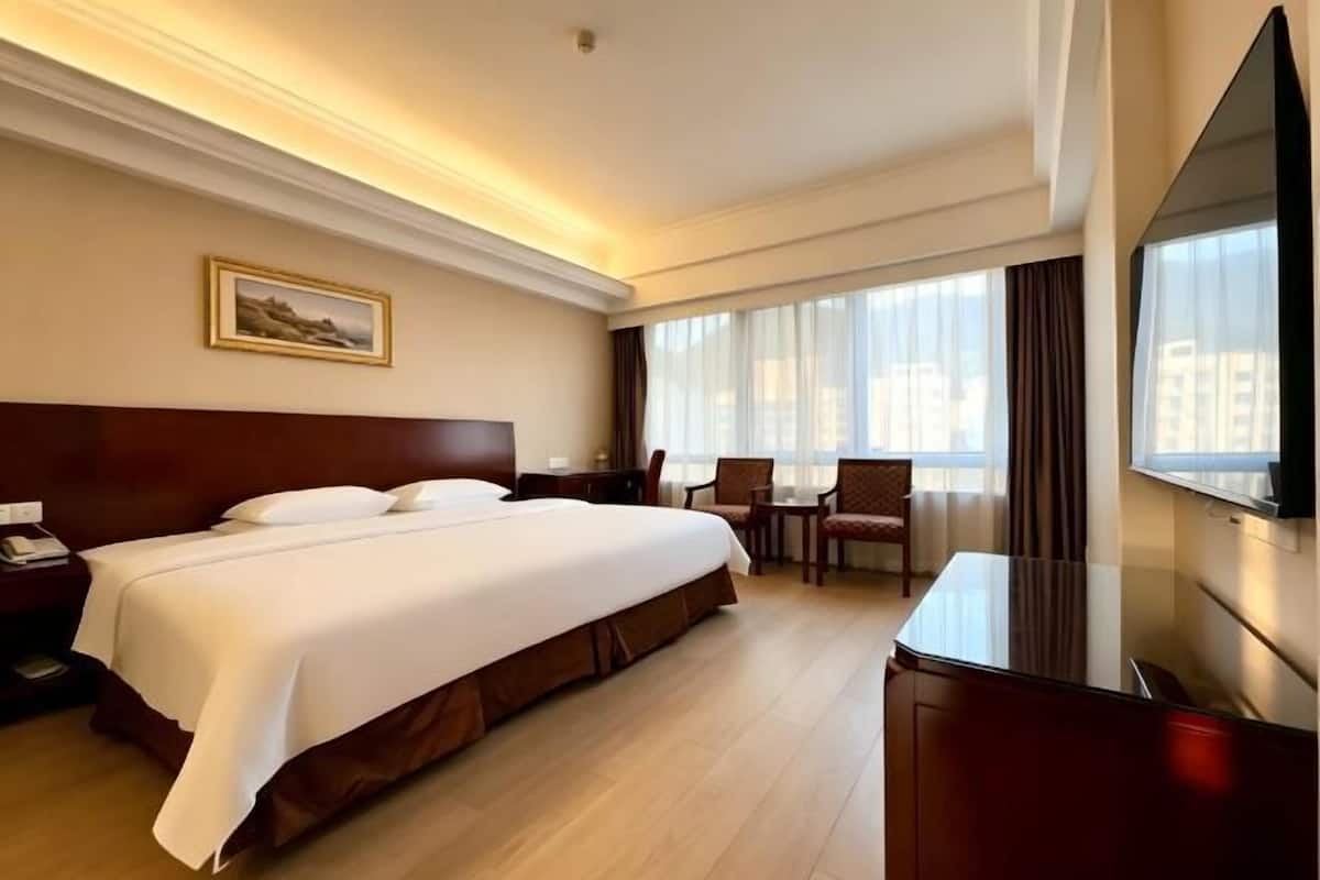 A hotel room with a large bed, a desk, a chair, a TV, and a window with curtains.