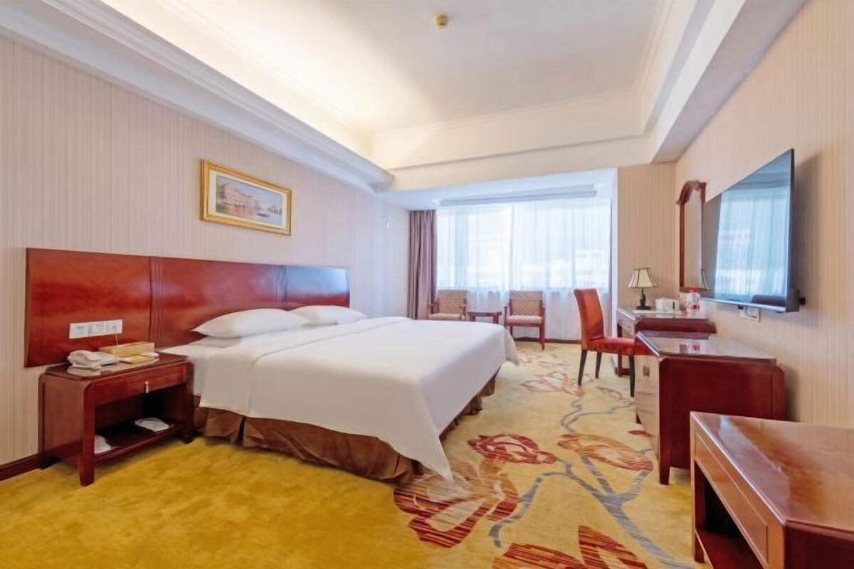 A hotel room with a large bed, a desk, a chair, a television, and a window with curtains.