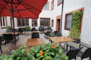 An outdoor seating area with tables and chairs, surrounded by a white building with windows and a red umbrella. - Bayerischer Hof Freising (Freising)