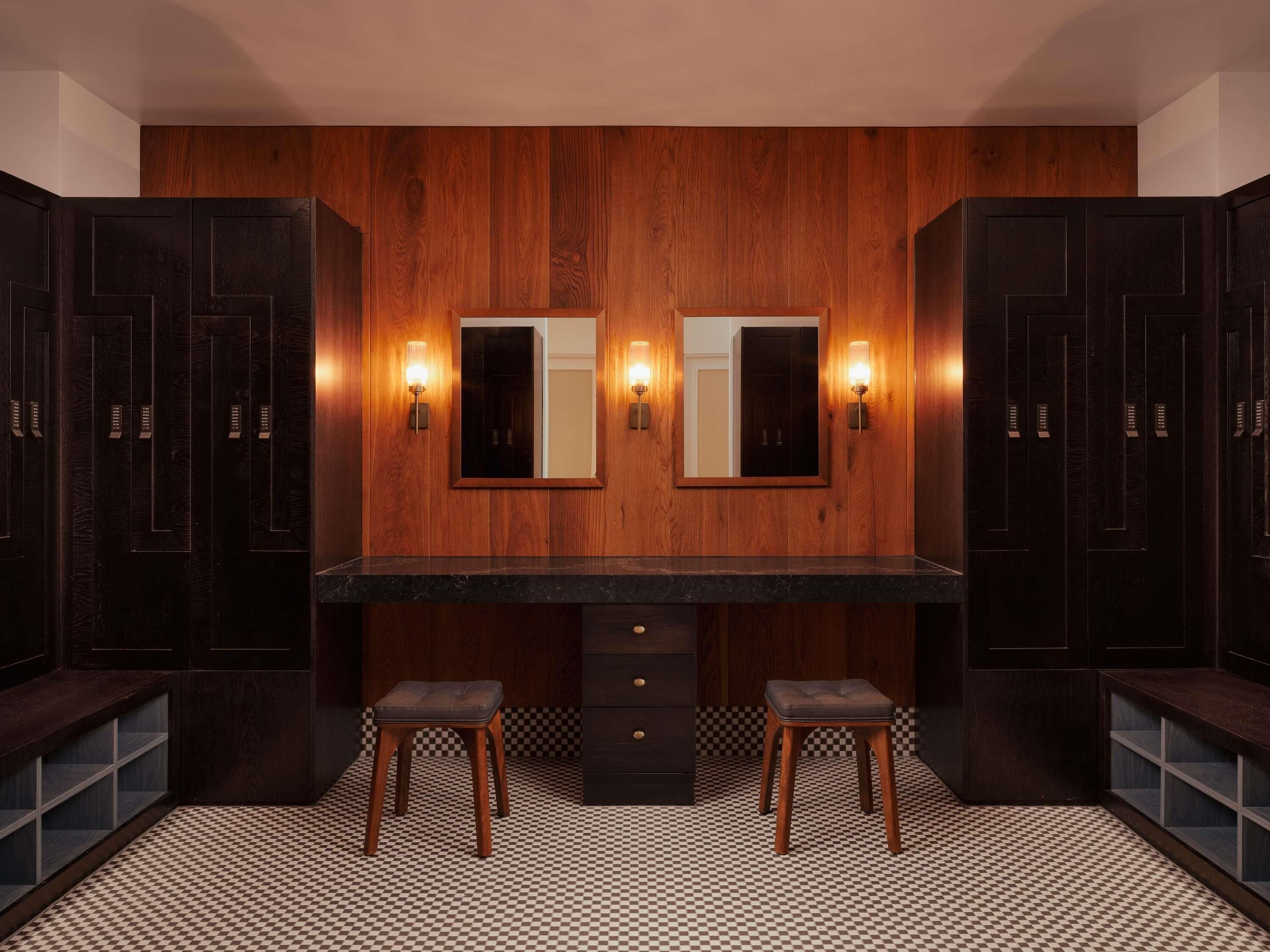 A modern bathroom with dark wood paneling, a central vanity with a black countertop, two stools, and wall-mounted mirrors with sconces.