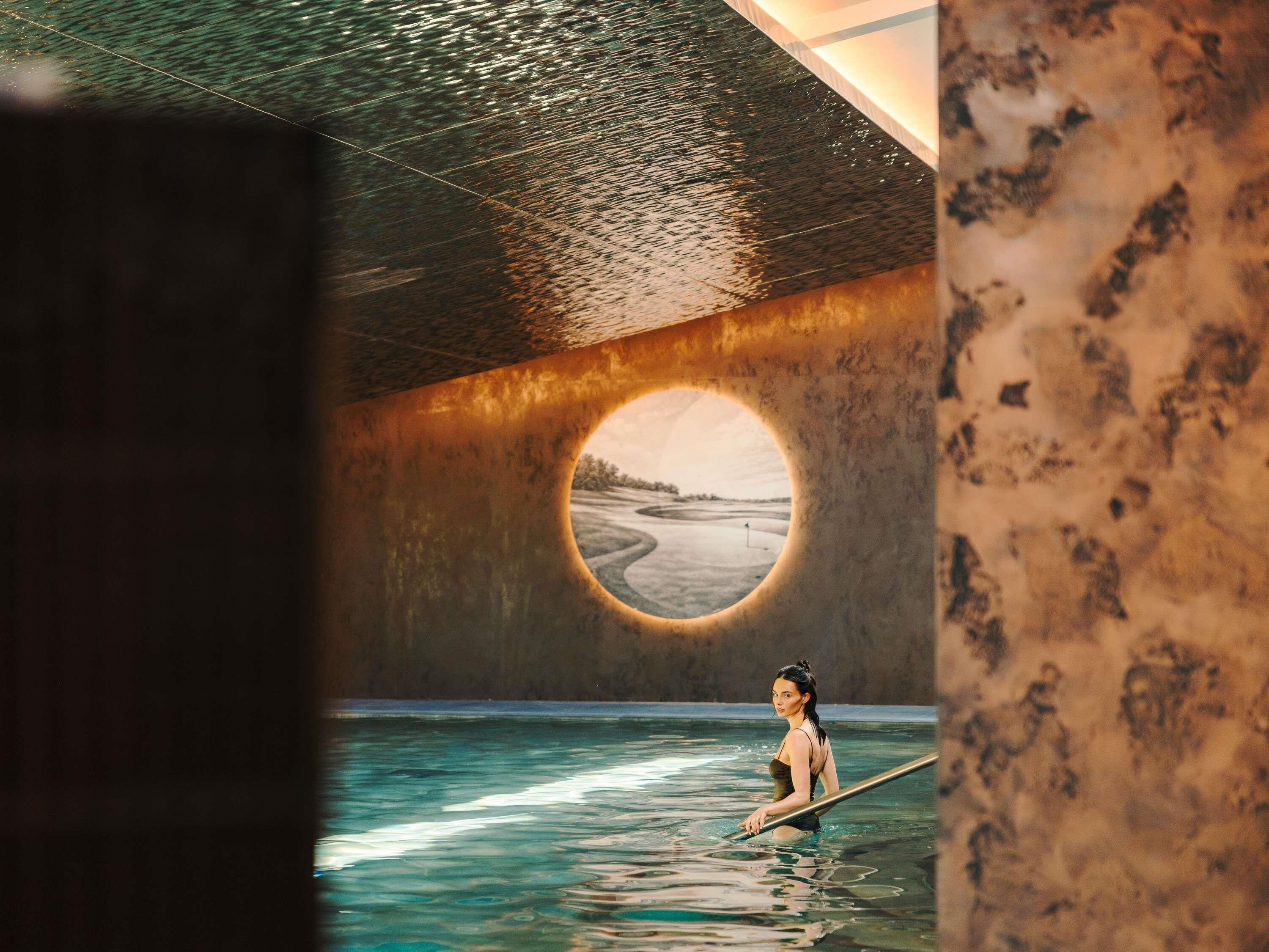 Una persona en una piscina con vista a una playa a través de un ventanal circular.