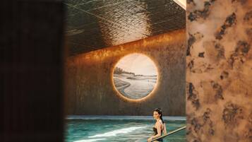 Una persona en una piscina con vista a una playa a través de un ventanal circular.
