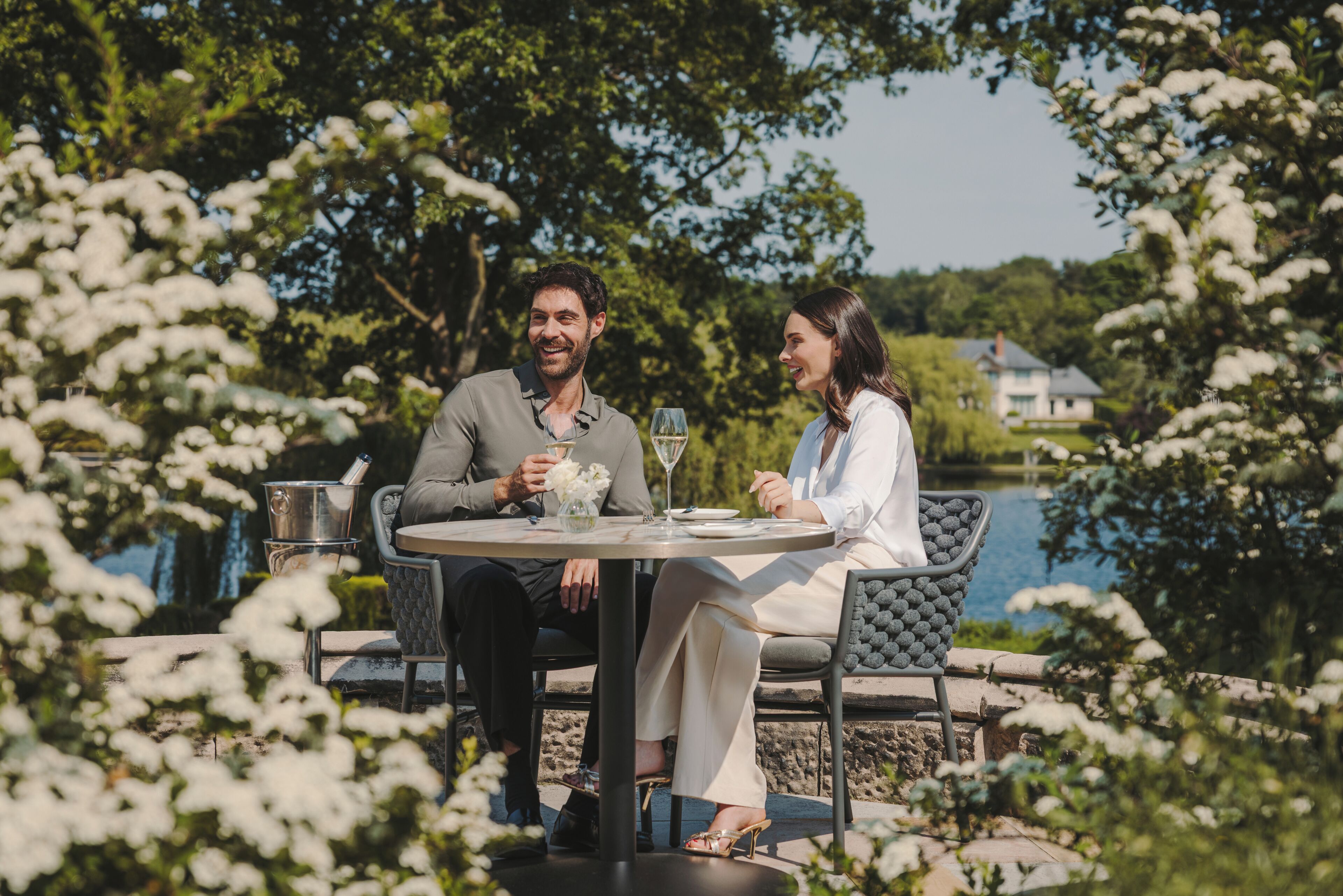 A couple dining at an outdoor table with a view of a lake and a house in the background.