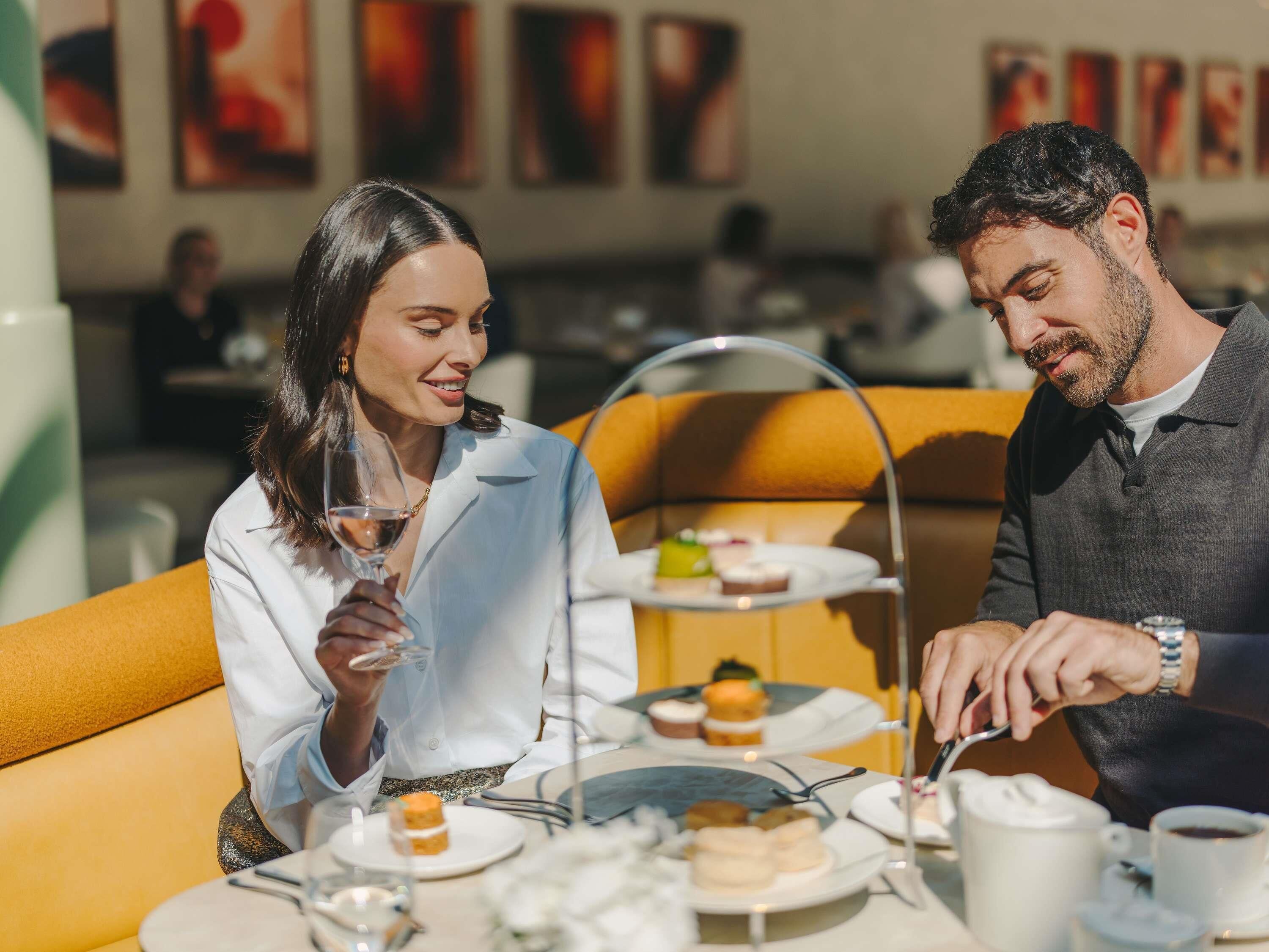 A couple dining at an outdoor café with a tiered tea service, a table set with plates, glasses, and a teapot.