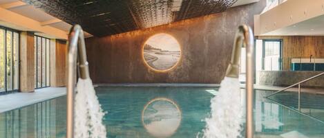 A swimming pool with a view of a beach through a circular window, surrounded by a modern interior with large windows and a high ceiling.