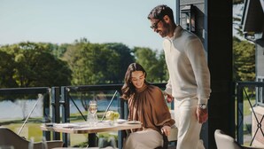 A dining table with chairs, a person standing, and a view of a body of water and trees. - Fairmont Cheshire The Mere (Knutsford)