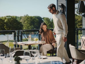 A dining table with chairs, a person standing, and a view of a body of water and trees. - Fairmont Cheshire The Mere (Opening July 2026) (Knutsford)