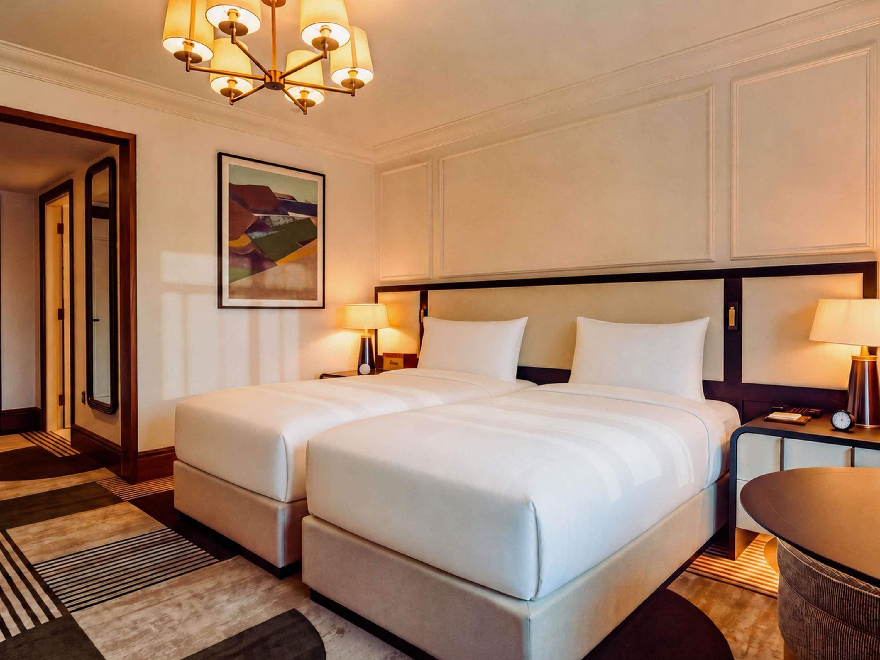 A hotel room with a double bed, two bedside tables with lamps, a large mirror, a painting on the wall, and a carpeted floor.