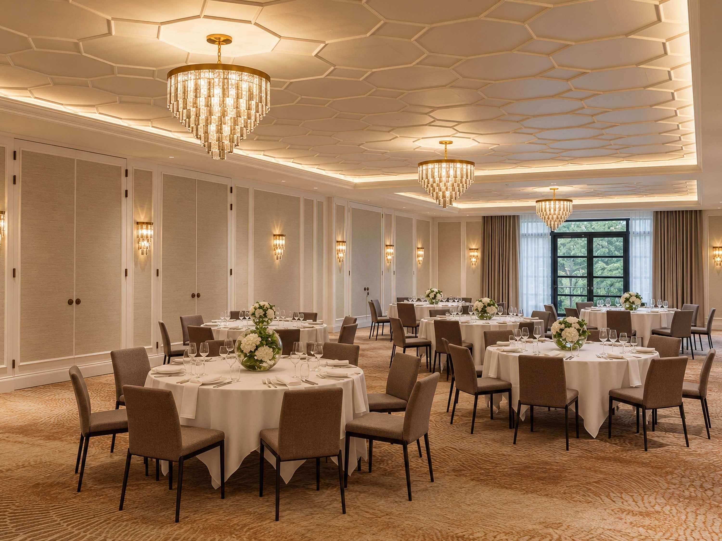 A spacious banquet hall with multiple round tables, elegant chandeliers, and a patterned ceiling.