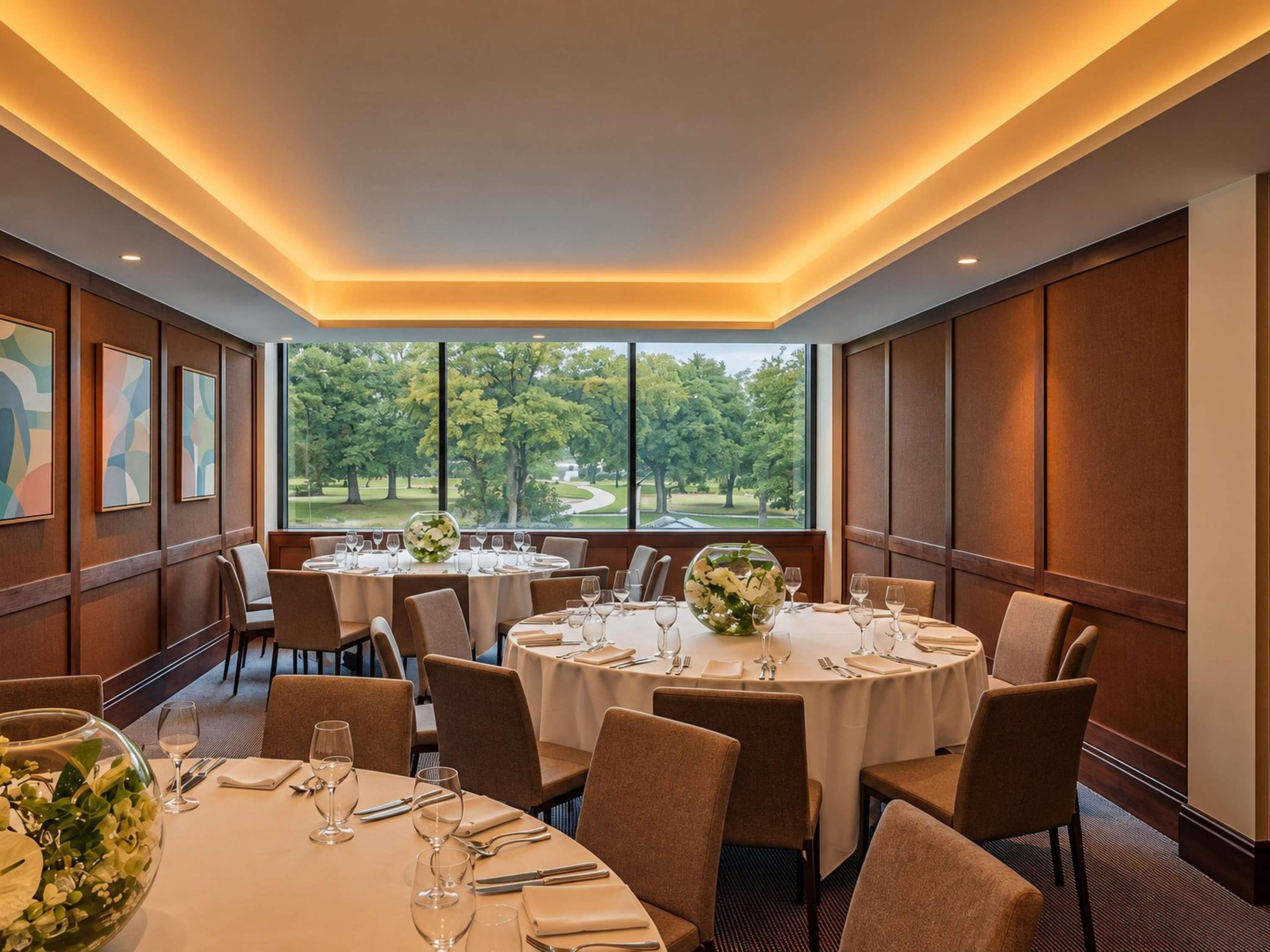 A dining area with round tables, chairs, and a view of trees through large windows.
