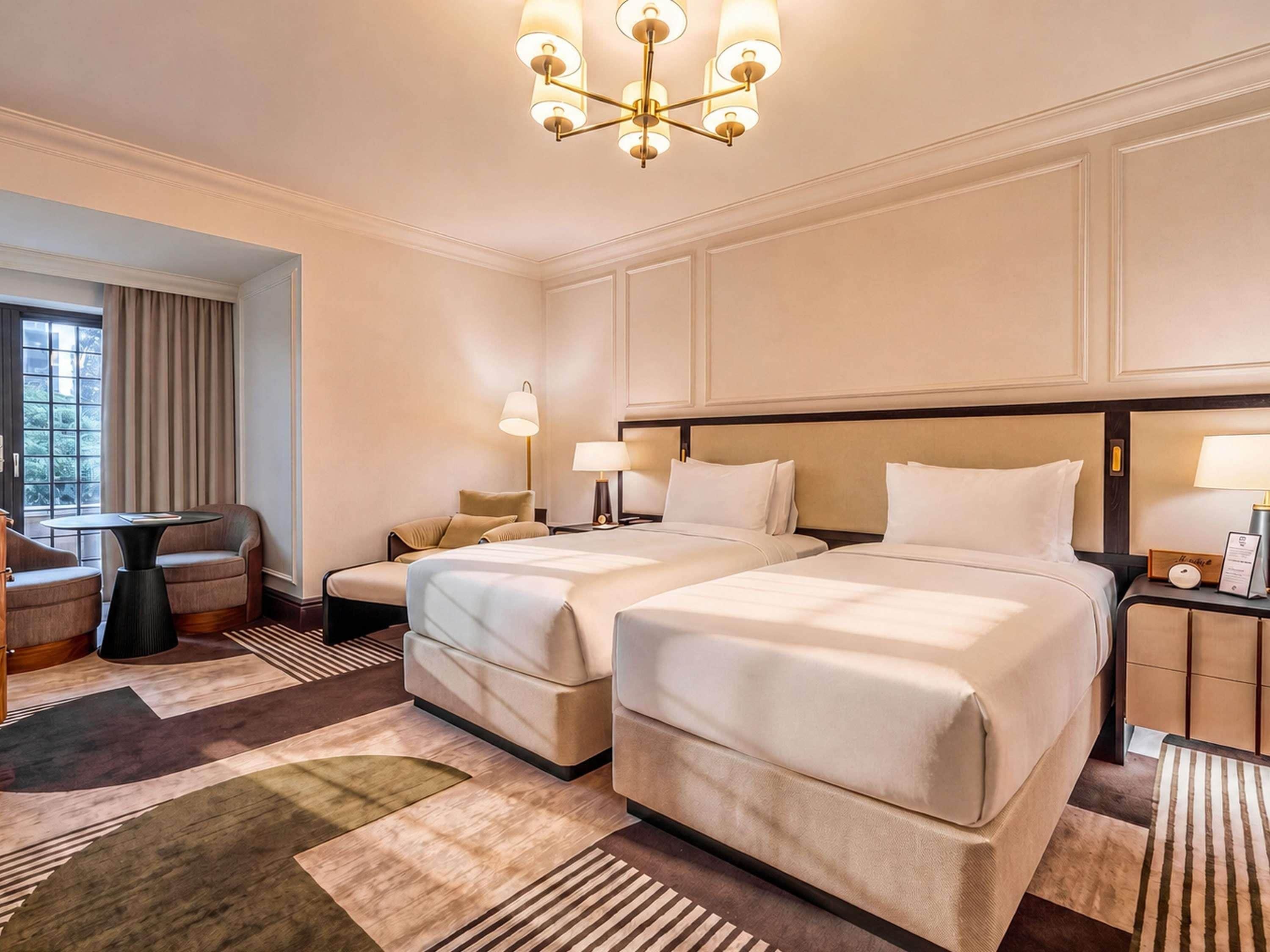 A hotel room with two beds, a sitting area with a chair and a table, a chandelier, and a window with curtains.