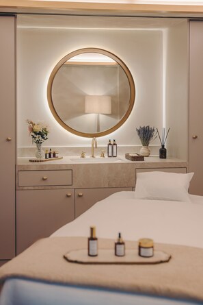 Une chambre d’hôtel avec un miroir rond, un lit, une table de chevet avec des articles de toilette et un vase rempli de fleurs.