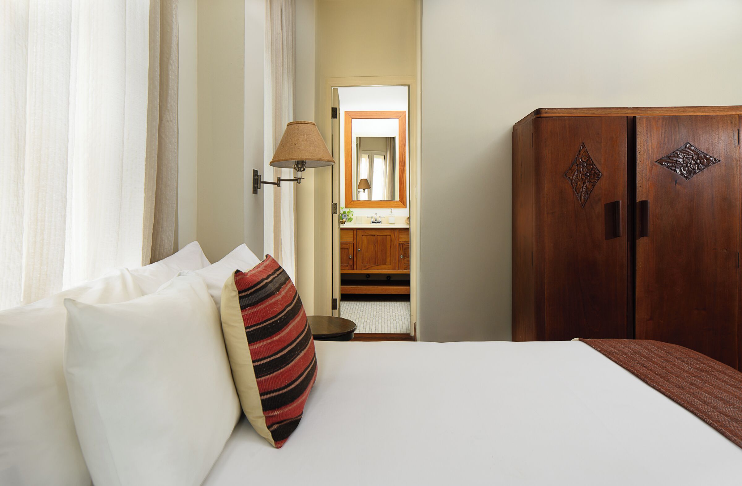 A hotel room with a bed, pillows, a lamp, a mirror, and a wooden wardrobe.