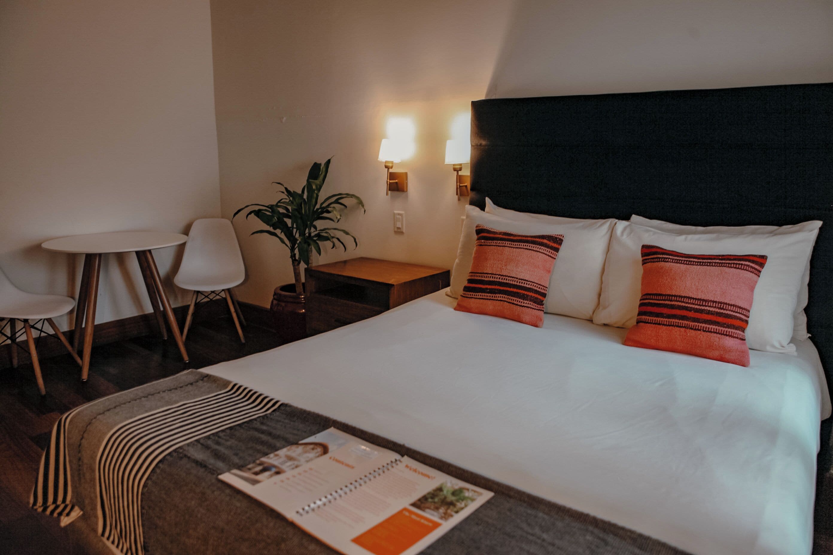 Ett hotellrum med en säng, ett litet bord, två stolar, en växt och väggmonterade lampor.