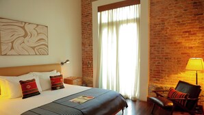 Ein Hotelzimmer mit Backsteinwand, einem großen Fenster mit Vorhängen, einem Bett mit Kissen, einem Nachttisch und einem Sessel.