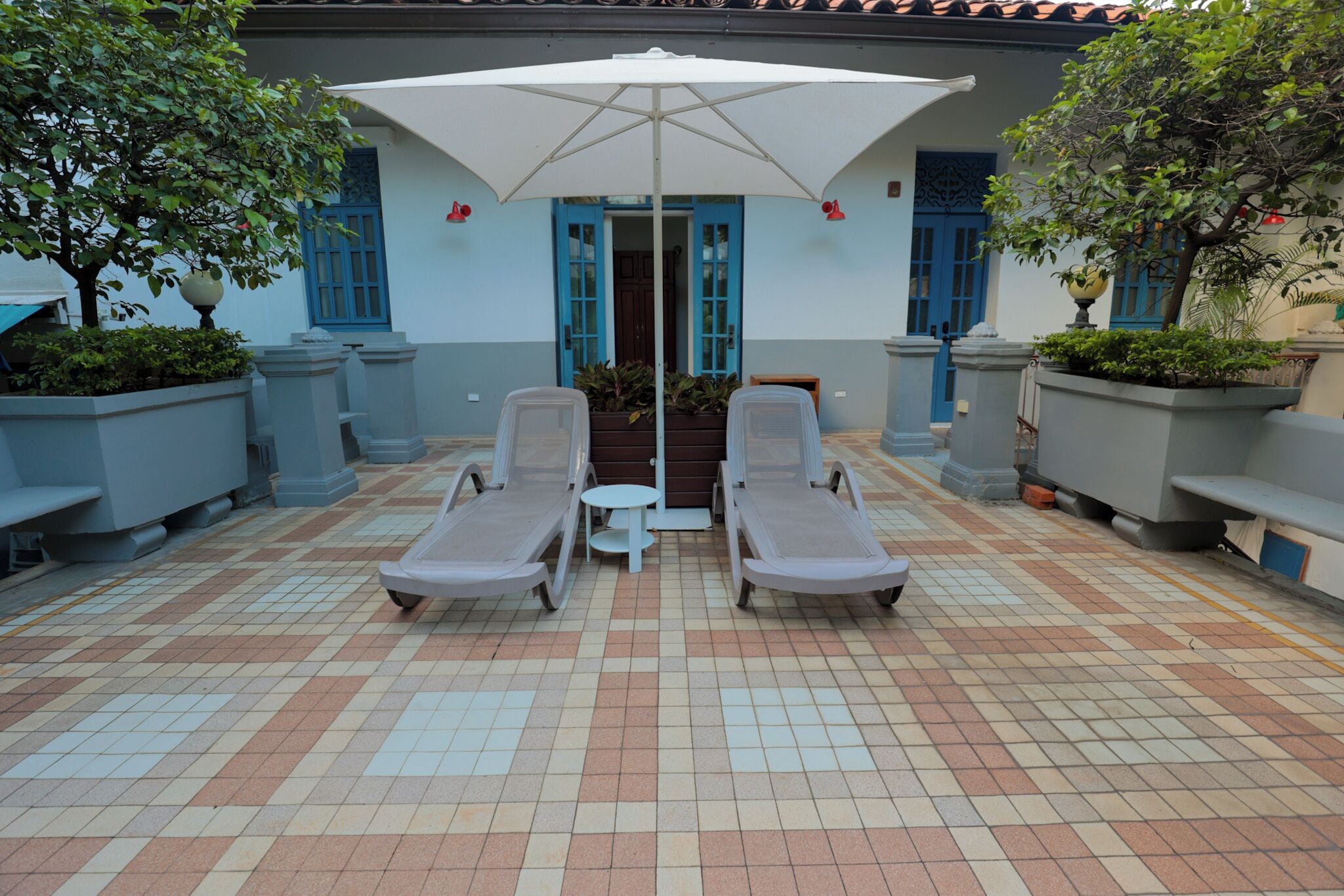 Un patio con baldosas, dos sillones, una mesa bajo un paraguas blanco, rodeado de macetas y un edificio con puertas y ventanas azules.