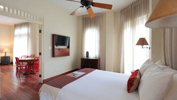 Une chambre d’hôtel comprenant un lit, un ventilateur de plafond, une télévision, une table à manger avec des chaises et une fenêtre avec des rideaux.