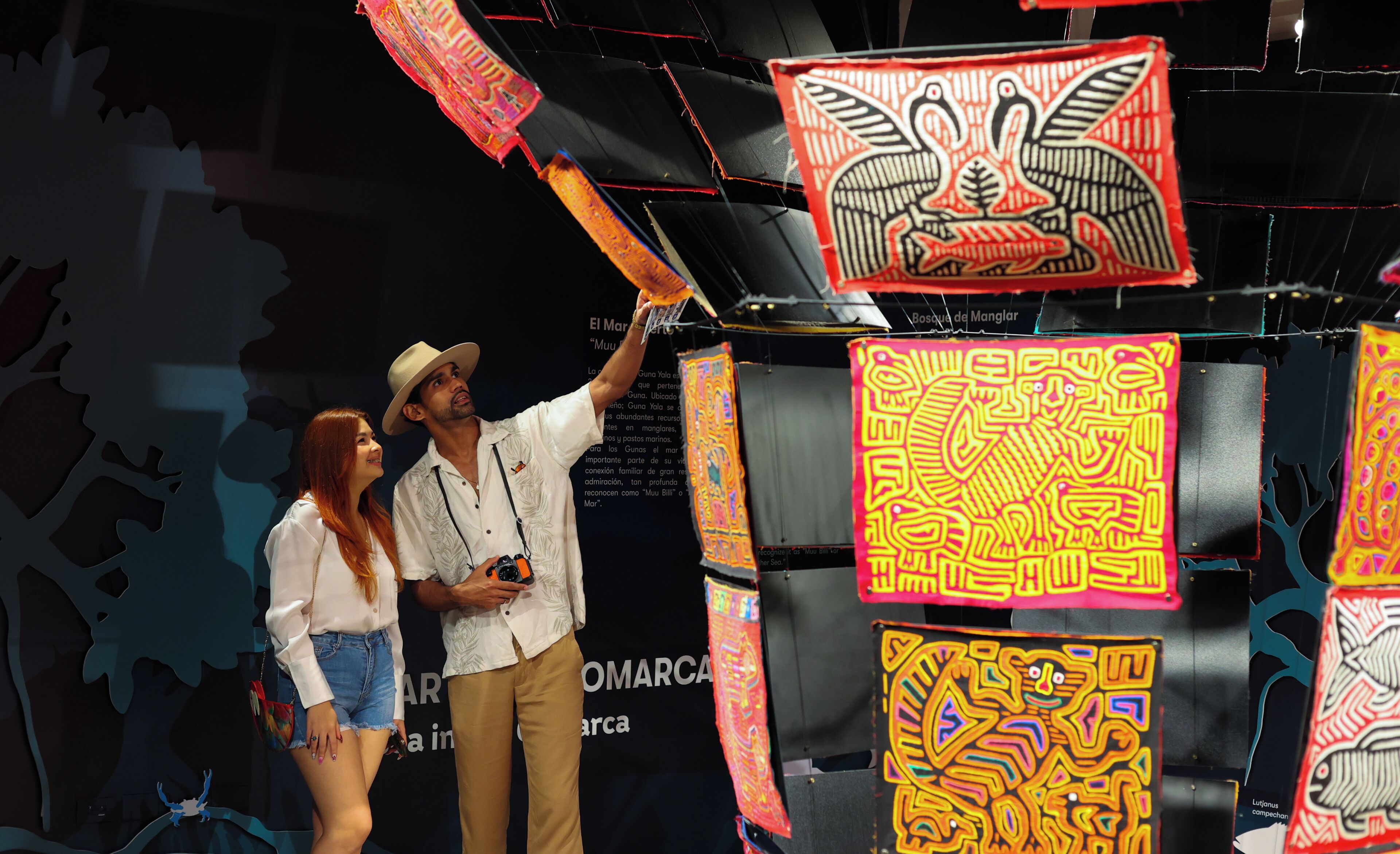 Un hombre y una mujer de pie frente a una exhibición de tapices coloridos sobre un fondo negro.