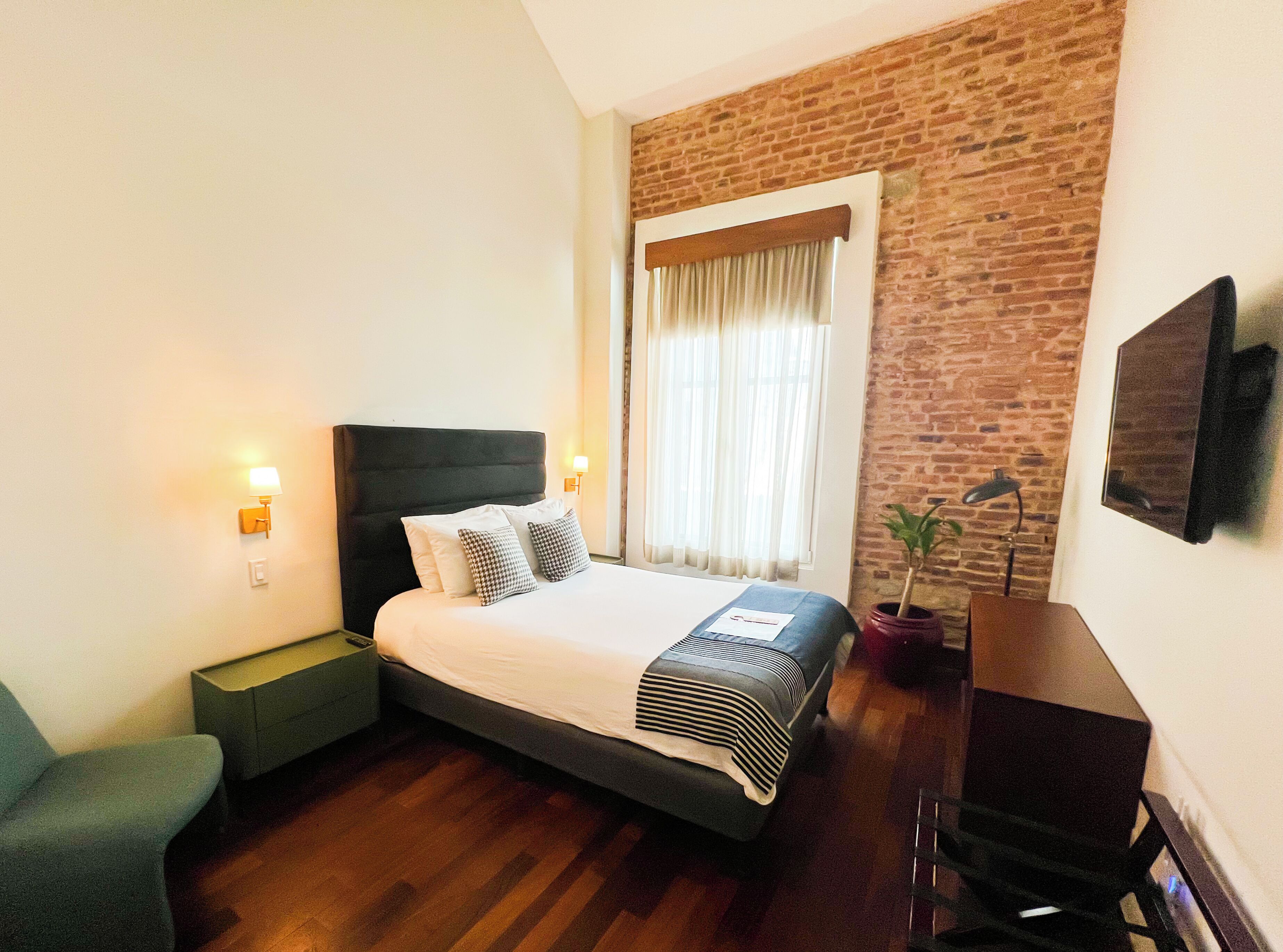 A hotel room with a brick wall, a bed with a headboard, bedside lamps, a desk with a chair, and a potted plant.