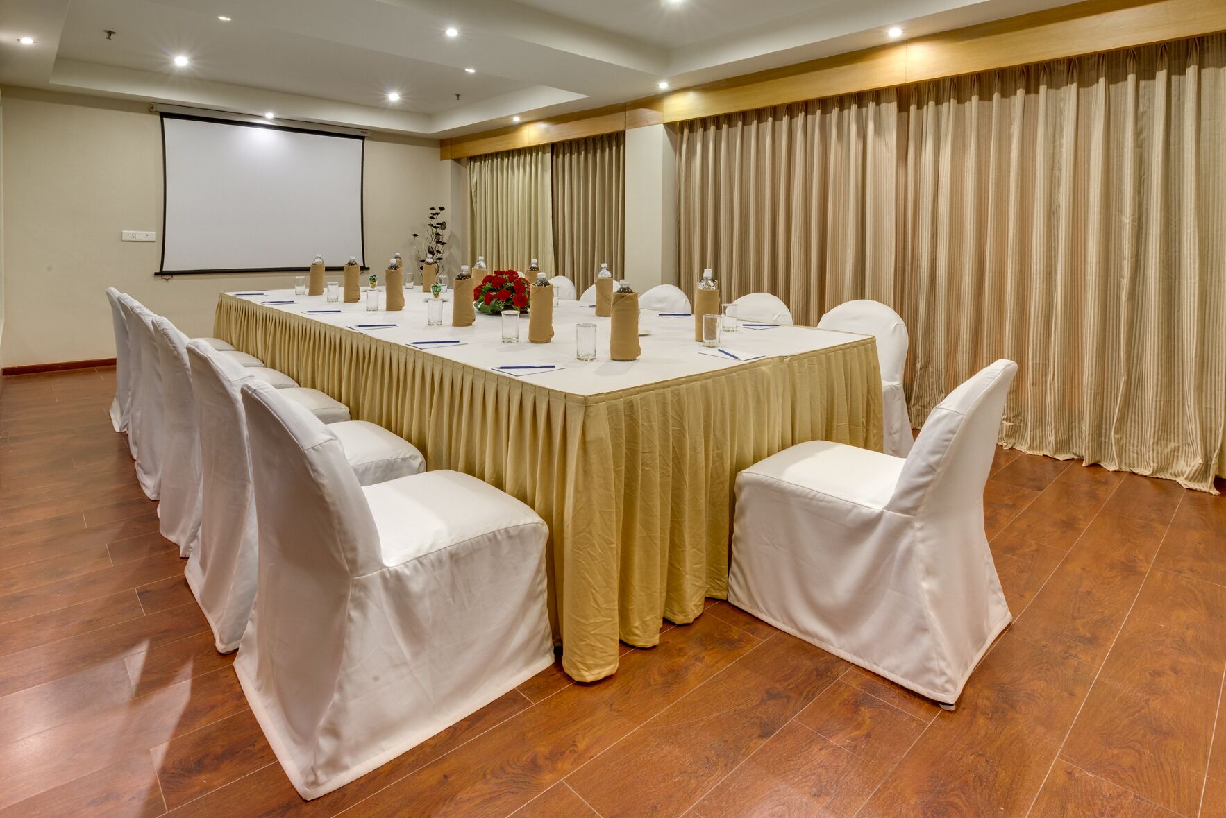 Una sala de conferencias con una mesa larga, sillas blancas y una pantalla para proyector.