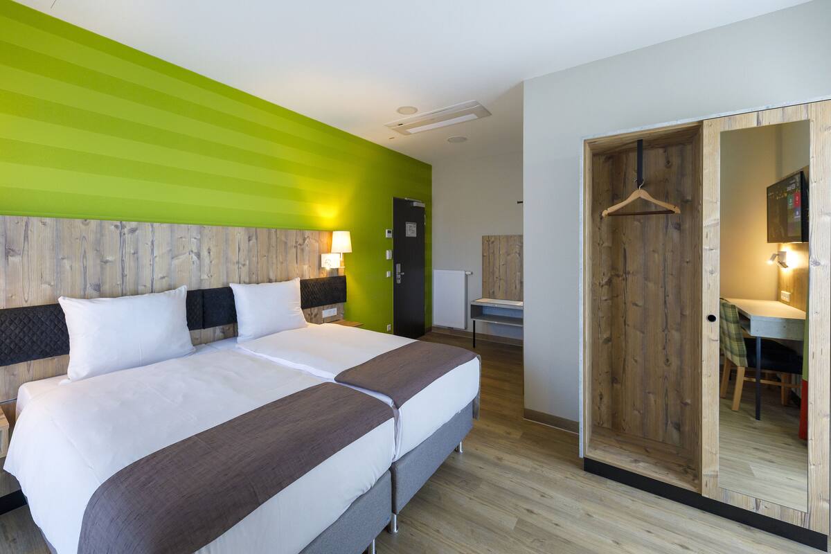 Quarto de hotel moderno com uma cama grande, cabeceira de madeira, parede verde decorativa, guarda-roupa de madeira e uma escrivaninha com cadeira.
