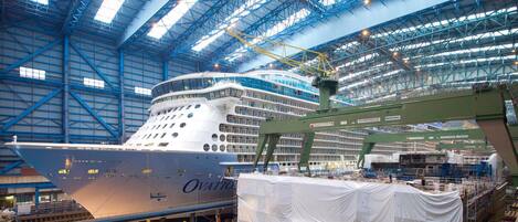 Un gran barco en construcción dentro de una amplia nave industrial con techos altos y vigas estructurales visibles.