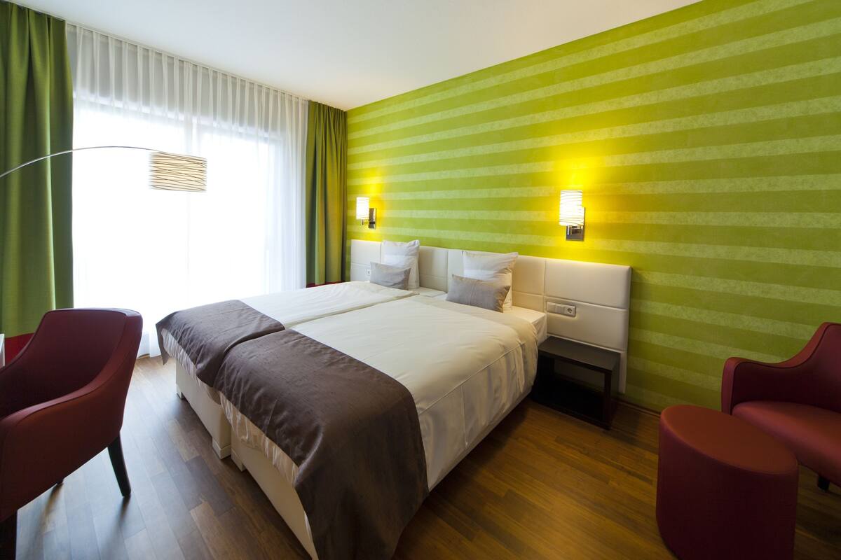 Quarto de hotel com uma cama grande, duas mesas de cabeceira, uma escrivaninha, duas cadeiras vermelhas e uma janela com cortinas.