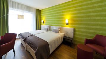 A hotel room with a large bed, two bedside tables, a nightstand, a desk, two red chairs, and a window with curtains.