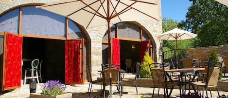Un patio al aire libre con suelo de piedra, macetas de madera con flores y mobiliario exterior con sombrillas.