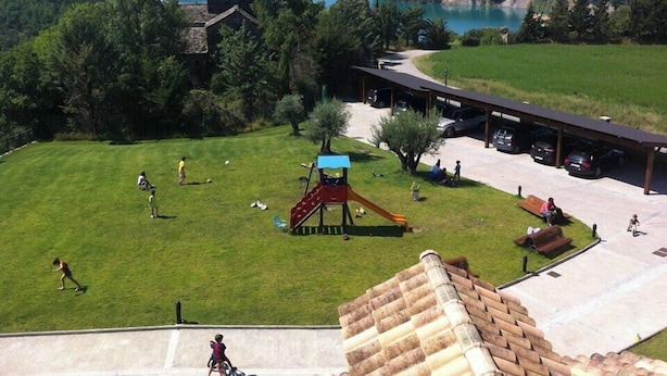 A playground with a slide and swings, a parking area, and a building with a sloped roof. - Casa Pirineo, Ainsa (Ainsa)