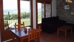 A room with a dining table, chairs, a sofa, a small table, and a view of a green landscape through large windows. - Casa Pirineo, Ainsa (Ainsa)