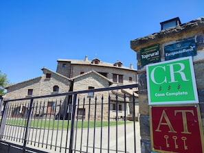 A stone building with a traditional architectural style, a metal gate, and signs indicating a fully equipped house and recommended accommodation.