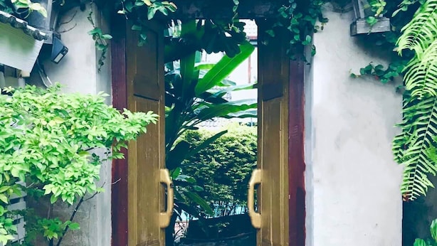 Una puerta de madera abierta que da a un estrecho sendero de piedra rodeado de abundante vegetación.