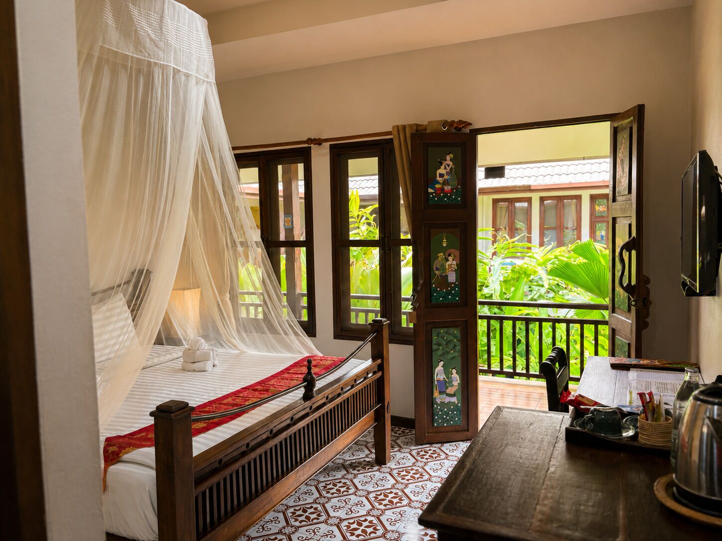A room with a bed, a table, and a balcony with plants.