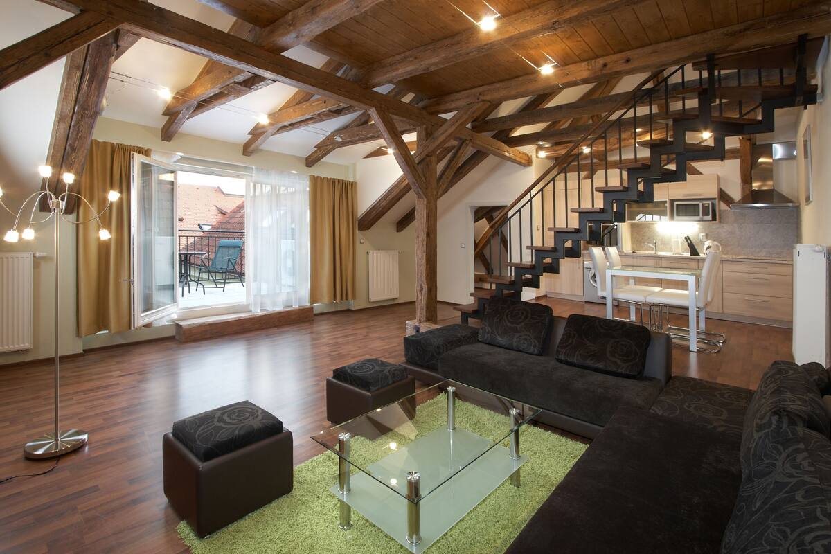 Un salón espacioso con una mesa de centro de cristal, sofás oscuros y una escalera que lleva a una cocina.
